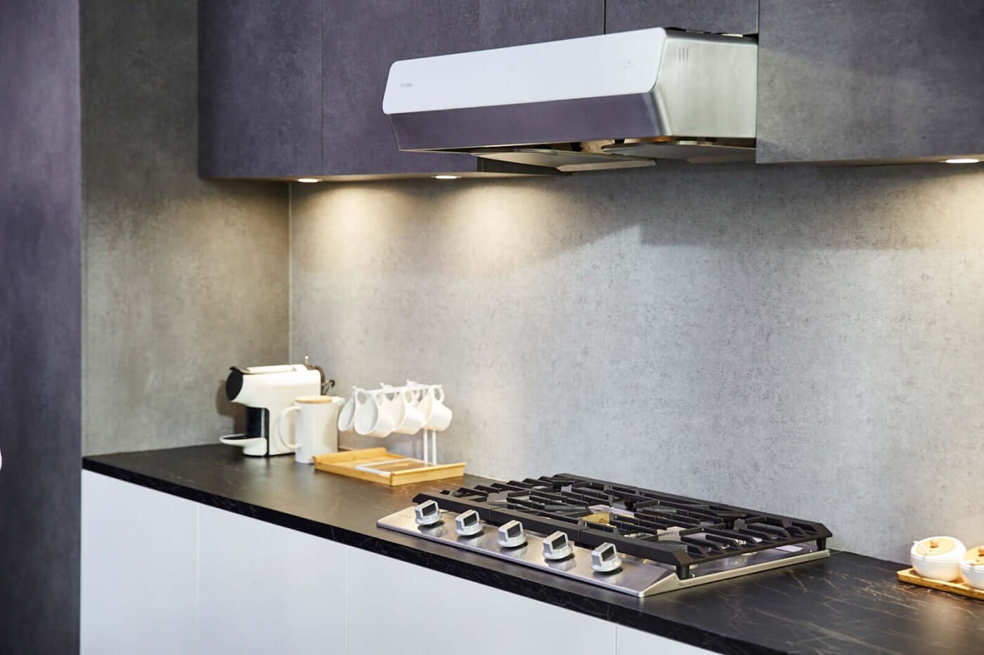Sleek modern kitchen corner showcasing a Fotile Pixie Air Series Slim Line under cabinet range hood above a gas cooktop, with a minimalist coffee maker, white cups on a wooden holder, and small candles adding a homey touch to the monochromatic decor.