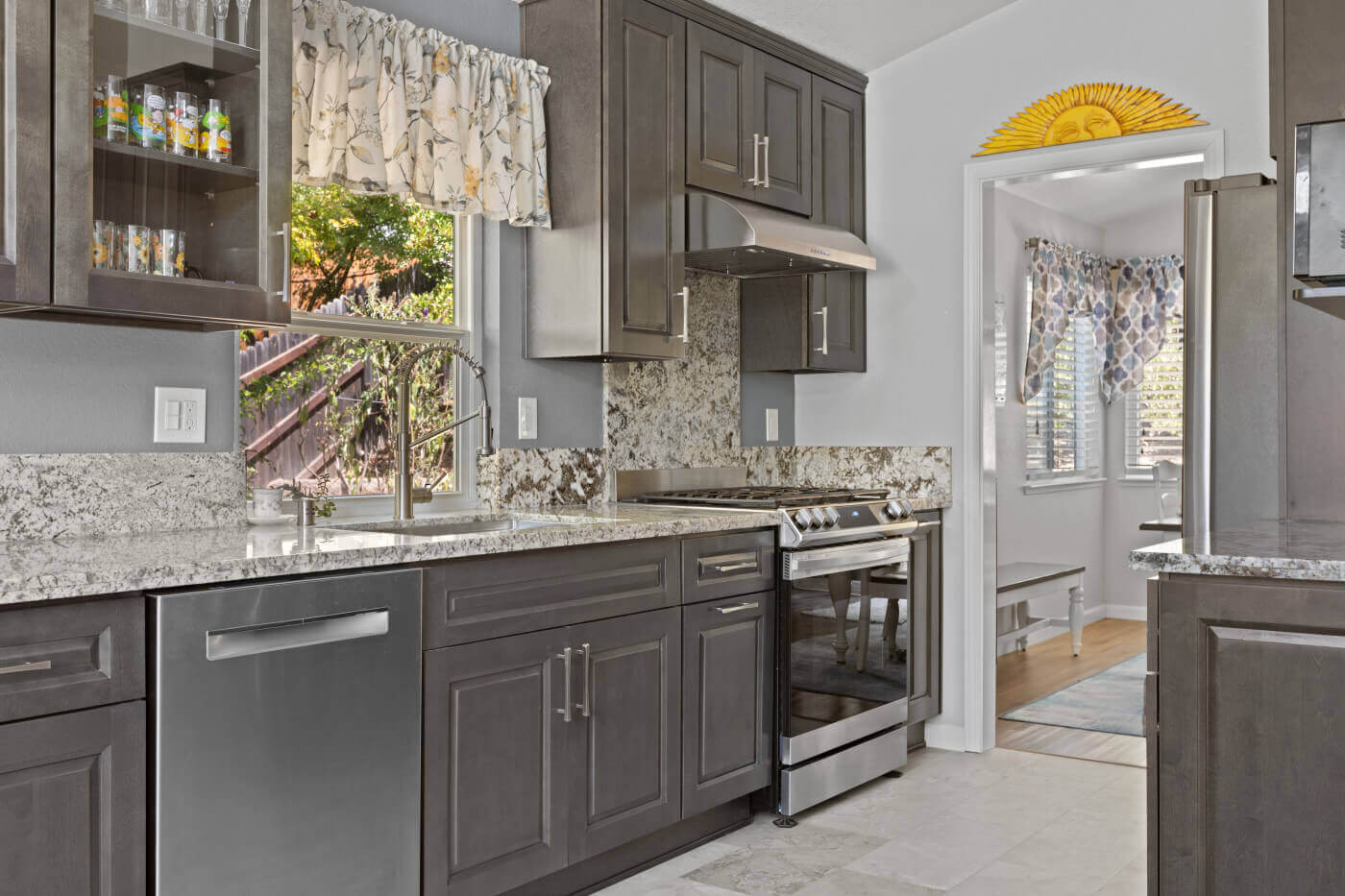 Modern kitchen interior with granite countertops and stainless steel appliances, framed by dark wood cabinetry and accented with a sunflower-themed wall decor above the doorway. The scene is brightened by natural light from a window adorned with bird-themed curtains.