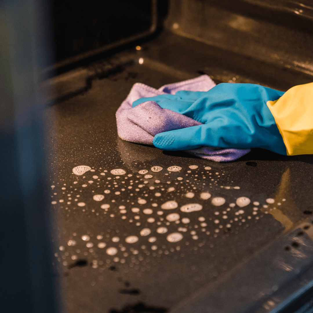 stock image of someone wiping up cleaner inside their oven with blue gloves on and a microfiber towel