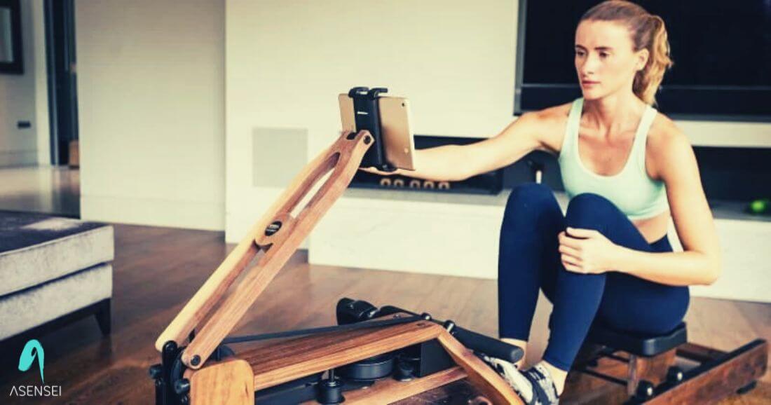 A woman sits on a rowing machine, in low light, wearing workout clothes. The asensei logo is in the corner