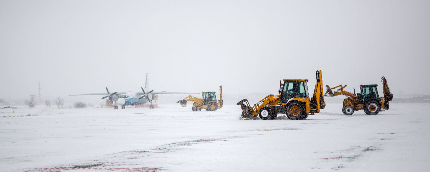 Airport snow removal vehicles during snowstorm with airplane in the background.